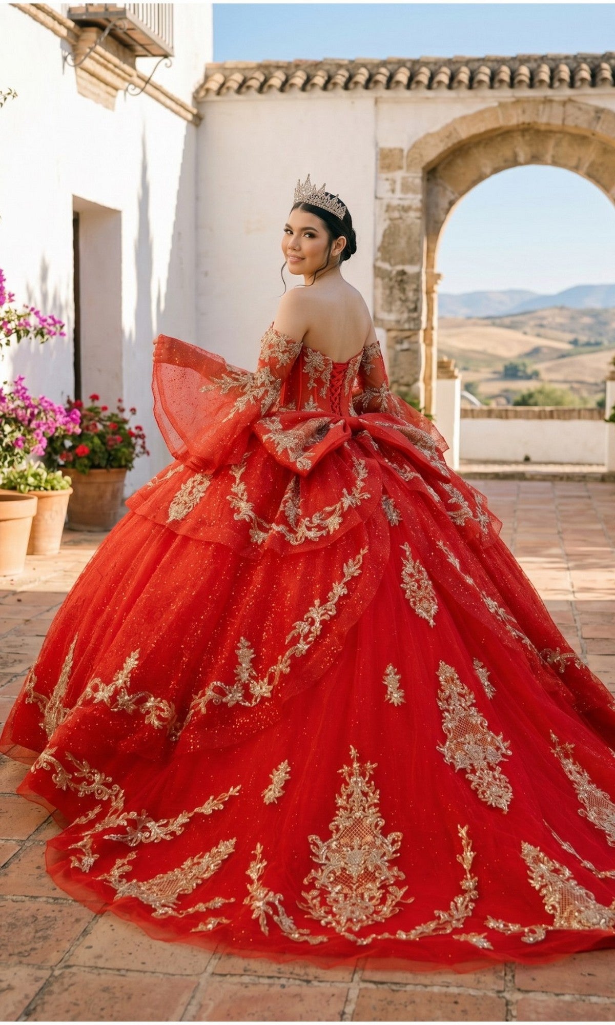 A young woman in the Quinceanera Dress 8575JQ By Cinderella Couture, adorned with red and gold embellishments and a sparkling tiara, stands outdoors on a tiled patio facing slightly away, with hills and a stone archway visible in the background.